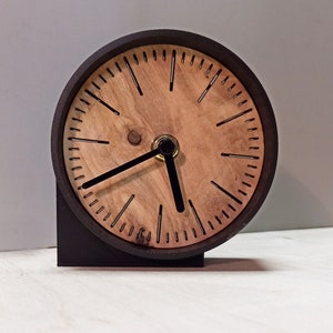 Könnte beinhalten: Eine schwarze und hölzerne Uhr mit einem einfachen Design. Das Zifferblatt der Uhr ist aus Holz und hat schwarze Stunden- und Minutenzeiger.