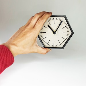 May include: A black hexagonal clock with a white face and black hands. The clock is being held by a hand wearing a red jumper.