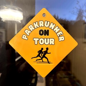 May include: A diamond-shaped yellow sign with a single hole at the top. The sign features the black text "PARKRUNNER ON TOUR" arched above the words "ON TOUR" in a bold font, with a black graphic of two people running below the text.