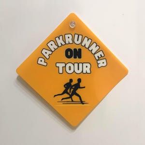 May include: A diamond-shaped yellow sign with a small clear fastener at the top. The sign features the black text PARKRUNNER ON TOUR in a bold font, accompanied by a black graphic illustration of two people running side by side.