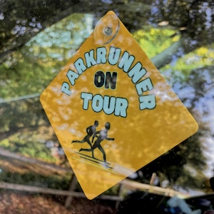May include: A diamond-shaped yellow car sign with a suction cup at the top. The sign features the text PARKRUNNER ON TOUR in light blue letters with black outlines, and a black graphic of two people running on a yellow background.