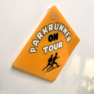 May include: A diamond-shaped yellow sign with a suction cup at the top. The sign features the black text "PARKRUNNER ON TOUR" in a bold font and a black graphic illustration of two people running, positioned below the text on the yellow surface.
