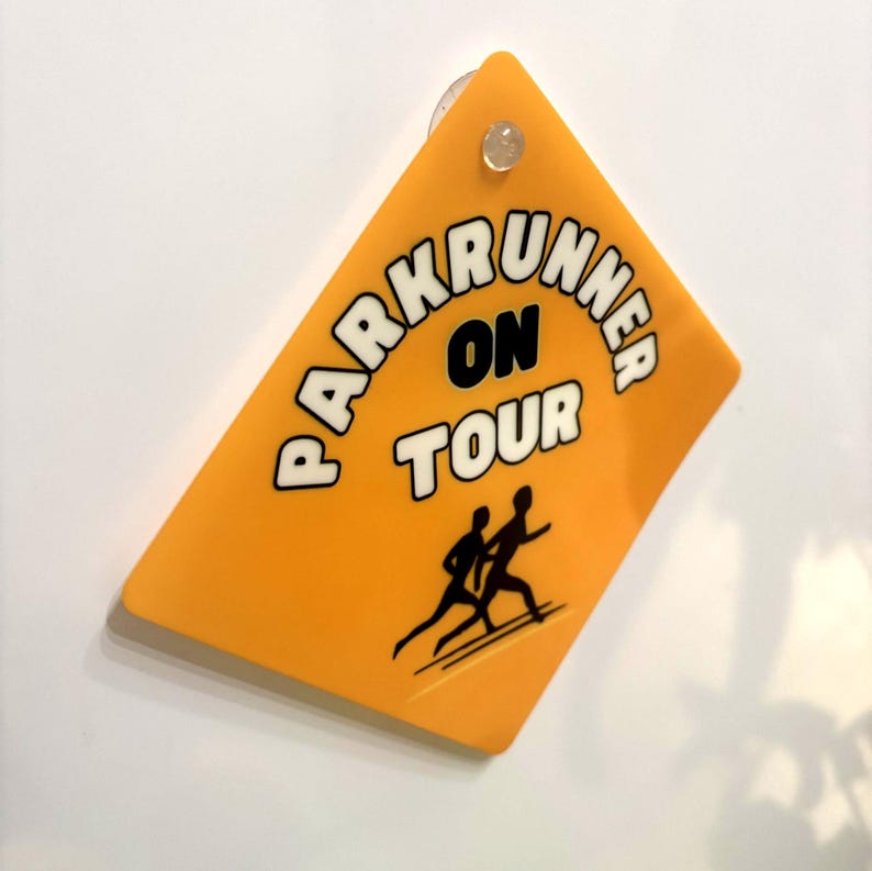 May include: A diamond-shaped yellow sign with a suction cup at the top. The sign features the black text "PARKRUNNER ON TOUR" in a bold font and a black graphic illustration of two people running, positioned below the text on the yellow surface.