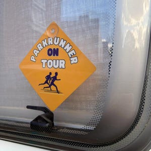 May include: A diamond-shaped yellow sign with a suction cup, featuring the text "PARKRUNNER ON TOUR" in dark blue letters and a graphic of two people running. The sign is attached to a glass window, displaying a clear warning for road safety.