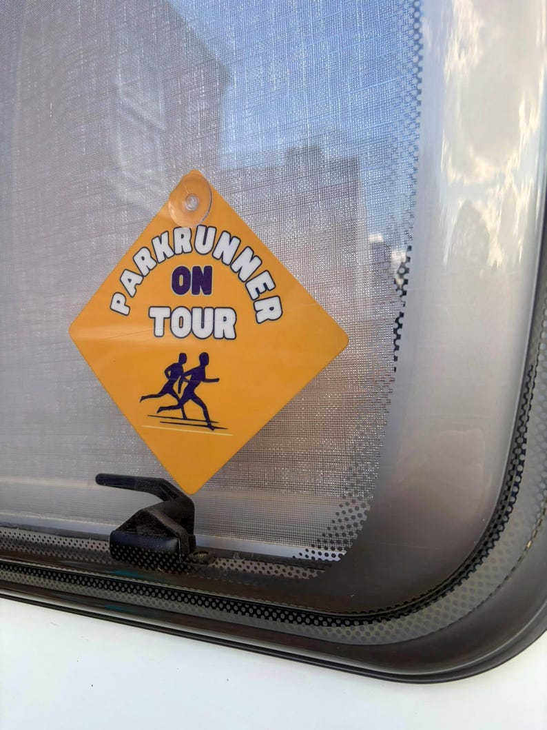 May include: A diamond-shaped yellow sign with a suction cup, featuring the text "PARKRUNNER ON TOUR" in dark blue letters and a graphic of two people running. The sign is attached to a glass window, displaying a clear warning for road safety.