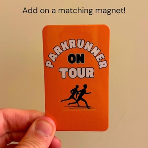 May include: A rectangular orange card featuring the black text PARKRUNNER ON TOUR and a black silhouette graphic of two people running. Above the card, the text reads Add on a matching magnet! The card has rounded corners and a smooth finish.