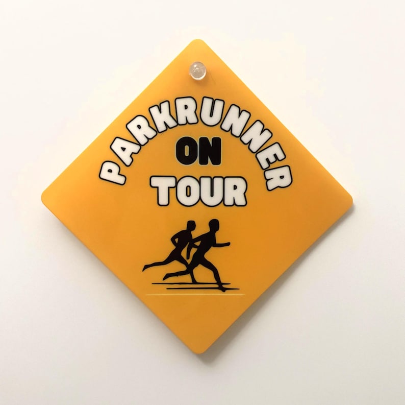 May include: A diamond-shaped yellow sign with a small clear pin at the top. The sign features the black text "PARKRUNNER ON TOUR" in a bold font and a black graphic of two people running, positioned below the text on a smooth surface.