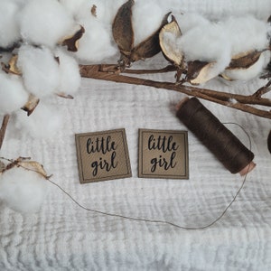 Könnte beinhalten: Zwei quadratische, beige Stoffetiketten mit den Worten "little girl" in schwarzer Schrift. Eine Spule braunen Fadens und ein Faden liegen neben den Etiketten. Der Hintergrund umfasst Baumwollkapseln und einen Zweig.