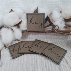 Könnte beinhalten: Vier quadratische, taupefarbene Etiketten mit dem Wort "hey" in schwarzer Schrift. Die Etiketten sind auf einer weißen Oberfläche mit Baumwolle und getrockneten Pflanzenelementen im Hintergrund angeordnet. Die Etiketten haben eine lederartige Textur.