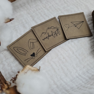 Könnte beinhalten: Drei braune Papierkarten mit schwarzen Tintenillustrationen. Die erste Karte zeigt ein Kaninchen-Gesicht mit einer Herznase. Die zweite Karte zeigt eine Bergkette mit einer Sonne und dem Wort "Wanderlust". Die dritte Karte zeigt ein Papierflugzeug mit einem Herzschwanz.