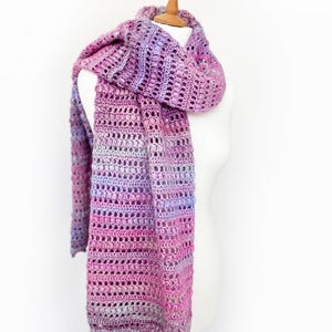 以下が含まれることがあります： マゼンタ、バイオレット、ラベンダー、ソフトピンクの色合いの段染め糸を使用した、長方形のクロッシェ編みのスカーフ。透かし編みと密なクラスター編みの列が交互に繰り返される、質感のあるステッチパターンが特徴です。