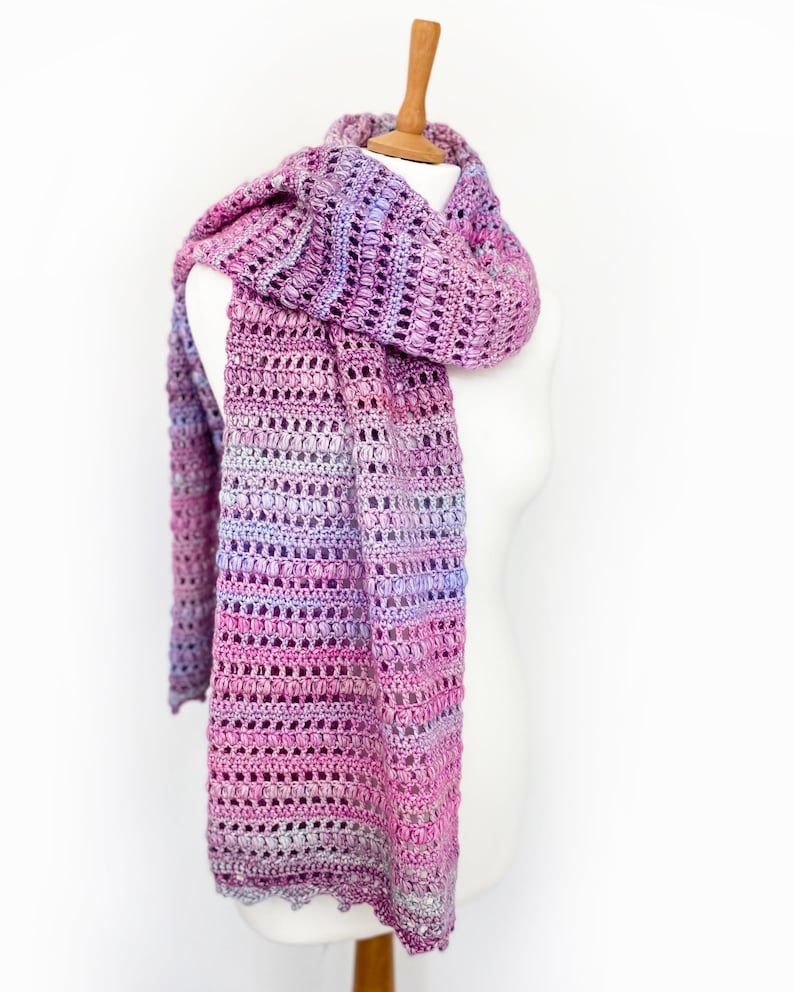 May include: A long, rectangular crochet scarf featuring a variegated yarn pattern in shades of magenta, violet, lavender, and soft pink. The scarf displays a repeating textured stitch pattern with alternating rows of openwork and dense clusters.