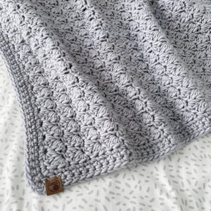 May include: A gray crocheted baby blanket with a textured pattern. The blanket has a small brown tag with the text "Loop Yarns" on it.