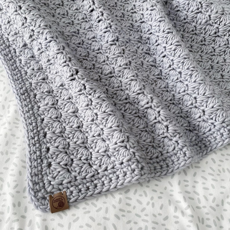 May include: A gray crocheted baby blanket with a textured pattern. The blanket has a small brown tag with the text "Loop Yarns" on it.