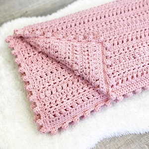 Pode incluir: Um cobertor de croch&ecirc; rosa claro dobrado, com um padr&atilde;o texturizado e uma borda ondulada. O cobertor &eacute; feito de fio macio e &eacute; dobrado sobre uma superf&iacute;cie branca e fofa. &Eacute; um item feito &agrave; m&atilde;o.