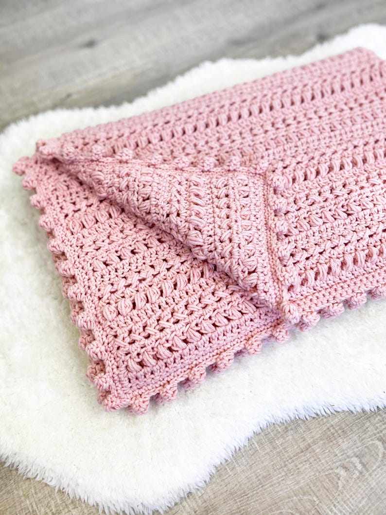 Pode incluir: Um cobertor de croch&ecirc; rosa claro dobrado, com um padr&atilde;o texturizado e uma borda ondulada. O cobertor &eacute; feito de fio macio e &eacute; dobrado sobre uma superf&iacute;cie branca e fofa. &Eacute; um item feito &agrave; m&atilde;o.