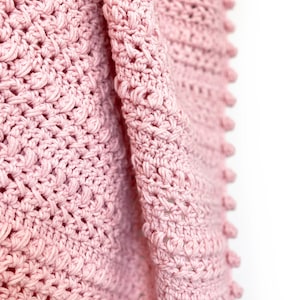 Pode incluir: Close-up de um cobertor rosa de croch&ecirc; macio. O cobertor apresenta um padr&atilde;o de ponto texturizado com bolinhas nas bordas. O fio &eacute; rosa pastel claro, criando uma est&eacute;tica aconchegante. O cobertor est&aacute; dobrado, mostrando os detalhes intrincados do artesanato. Medidas em cent&iacute;metros.