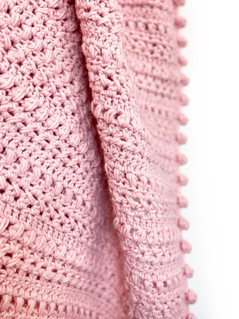 Pode incluir: Close-up de um cobertor rosa de croch&ecirc; macio. O cobertor apresenta um padr&atilde;o de ponto texturizado com bolinhas nas bordas. O fio &eacute; rosa pastel claro, criando uma est&eacute;tica aconchegante. O cobertor est&aacute; dobrado, mostrando os detalhes intrincados do artesanato. Medidas em cent&iacute;metros.