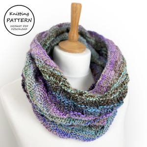 May include: A knitted cowl in shades of purple, blue, and green. The cowl is made with a textured stitch pattern and is displayed on a wooden mannequin.