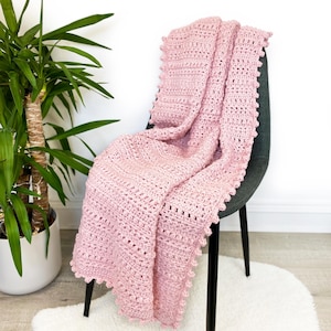 Pode incluir: Um cobertor de croch&ecirc; rosa claro drapeado sobre uma cadeira cinza escuro. O cobertor tem um padr&atilde;o texturizado e uma borda ondulada. Uma planta em vaso &eacute; vis&iacute;vel &agrave; esquerda, e um tapete branco e fofo est&aacute; no ch&atilde;o.