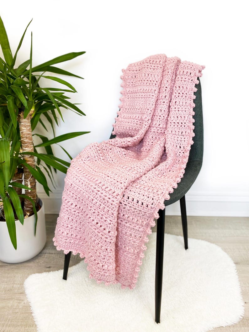 Pode incluir: Um cobertor de croch&ecirc; rosa claro drapeado sobre uma cadeira cinza escuro. O cobertor tem um padr&atilde;o texturizado e uma borda ondulada. Uma planta em vaso &eacute; vis&iacute;vel &agrave; esquerda, e um tapete branco e fofo est&aacute; no ch&atilde;o.