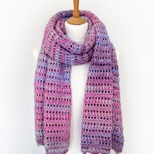 May include: A long, rectangular crochet scarf featuring a variegated yarn pattern in shades of magenta, purple, lavender, and light blue. The scarf displays a repeating textured stitch pattern with openwork sections and a scalloped bottom edge.