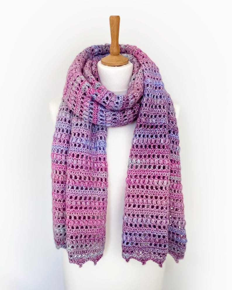 May include: A long, rectangular crochet scarf featuring a variegated yarn pattern in shades of magenta, purple, lavender, and light blue. The scarf displays a repeating textured stitch pattern with openwork sections and a scalloped bottom edge.