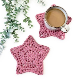 Puede incluir: Dos posavasos rosas en forma de estrella hechos a ganchillo. Uno de los posavasos sostiene una taza llena de una bebida marrón claro. Los posavasos están hechos de un hilo grueso y texturizado. Ramitas de hojas verdes en el fondo.