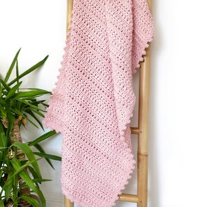 Pode incluir: Um cobertor de croch&ecirc; rosa claro macio drapeado sobre uma escada de madeira. O cobertor apresenta um padr&atilde;o de ponto texturizado e uma borda ondulada. &Eacute; um item aconchegante e feito &agrave; m&atilde;o.