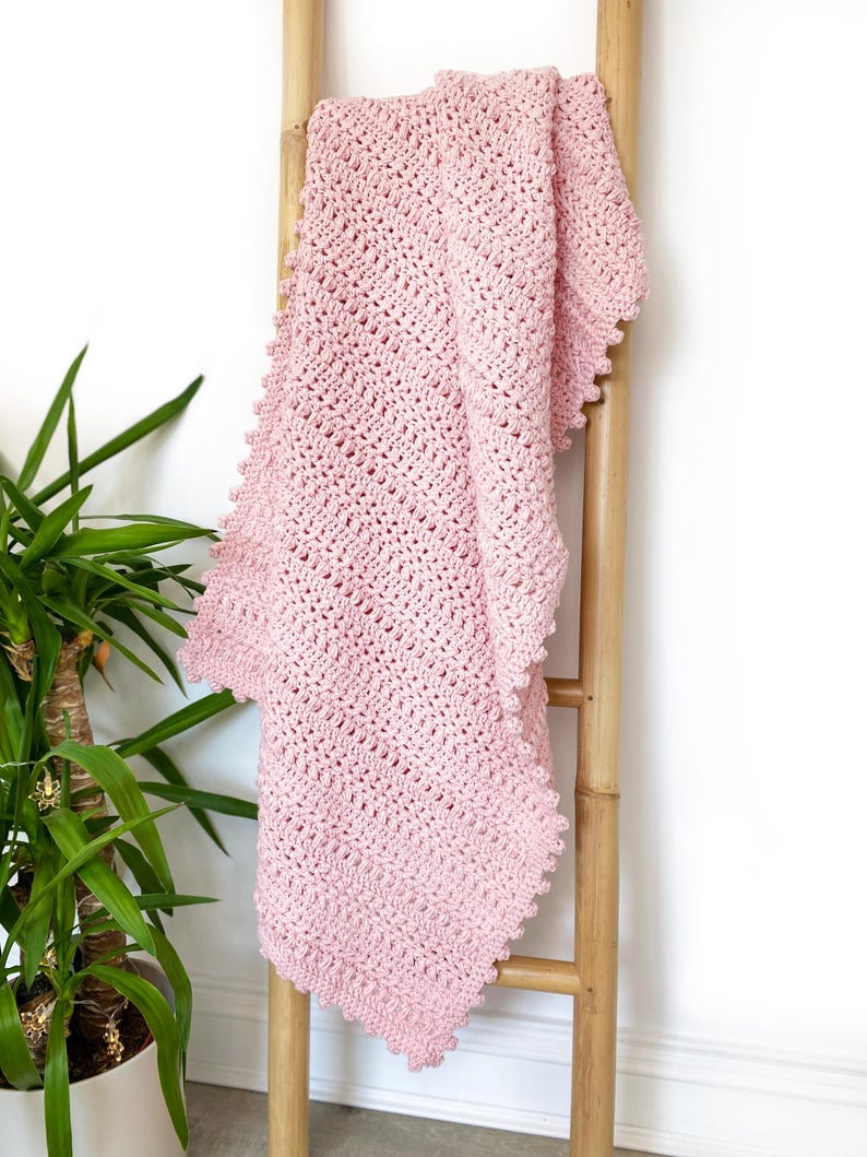 Pode incluir: Um cobertor de croch&ecirc; rosa claro macio drapeado sobre uma escada de madeira. O cobertor apresenta um padr&atilde;o de ponto texturizado e uma borda ondulada. &Eacute; um item aconchegante e feito &agrave; m&atilde;o.