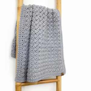 May include: A gray crocheted baby blanket hanging on a wooden ladder. The blanket has a delicate, lacy pattern.
