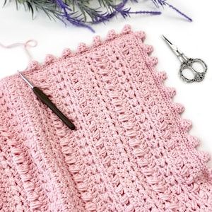 Pode incluir: Um cobertor de croch&ecirc; rosa claro feito &agrave; m&atilde;o, com um padr&atilde;o de ponto texturizado e uma borda ondulada. Uma agulha de croch&ecirc; repousa sobre o cobertor, com uma tesoura decorativa e galhos de flores roxas por perto. O cobertor &eacute; feito de fio macio.