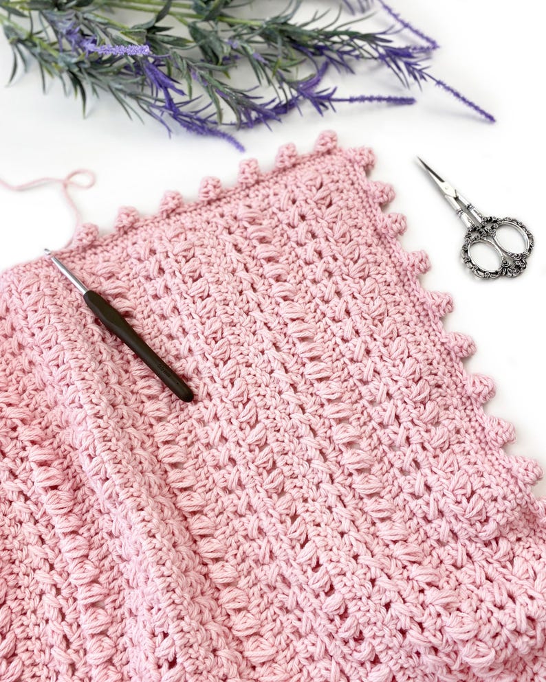Pode incluir: Um cobertor de croch&ecirc; rosa claro feito &agrave; m&atilde;o, com um padr&atilde;o de ponto texturizado e uma borda ondulada. Uma agulha de croch&ecirc; repousa sobre o cobertor, com uma tesoura decorativa e galhos de flores roxas por perto. O cobertor &eacute; feito de fio macio.
