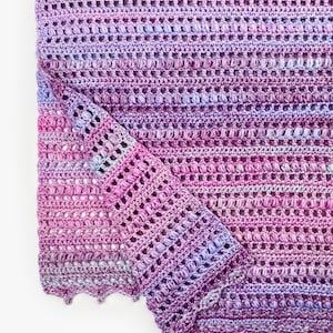 May include: A hand-crocheted blanket featuring a variegated yarn pattern in shades of magenta, violet, and light blue. The textile displays a repeating texture of horizontal rows of puff stitches and openwork, finished with a scalloped edge.