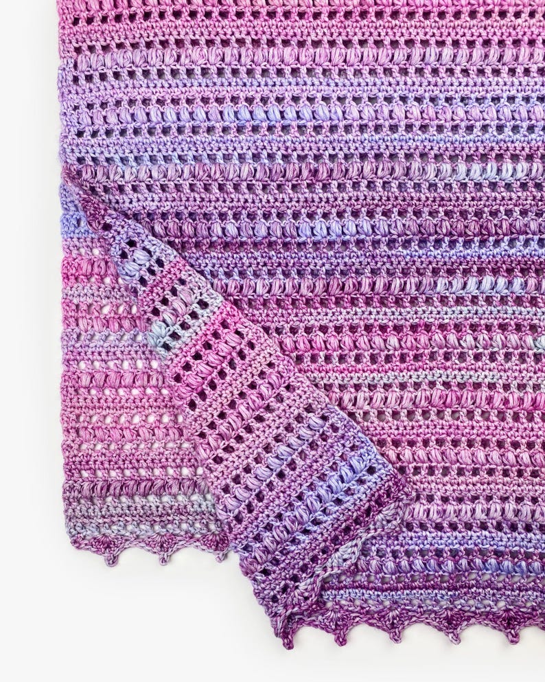 May include: A hand-crocheted blanket featuring a variegated yarn pattern in shades of magenta, violet, and light blue. The textile displays a repeating texture of horizontal rows of puff stitches and openwork, finished with a scalloped edge.