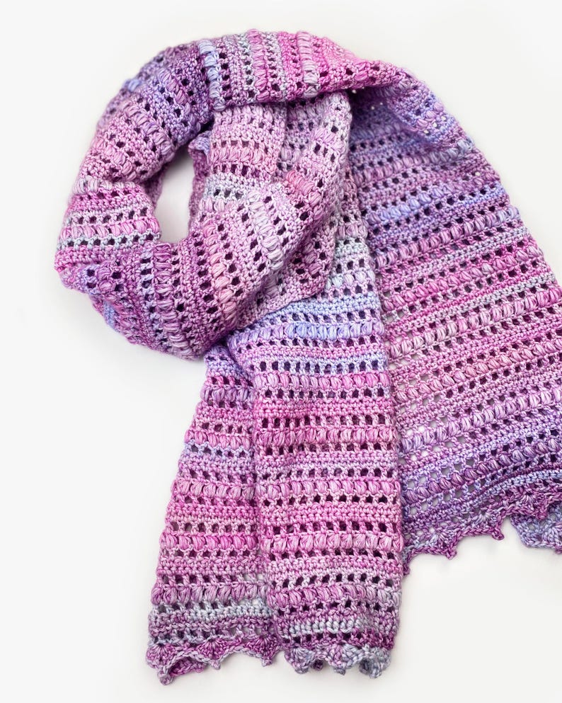 May include: A hand-crocheted scarf featuring a variegated yarn pattern in shades of magenta, lavender, and soft purple. The accessory displays a textured stitch design with alternating rows of openwork and dense clusters, finished with a scalloped edge.