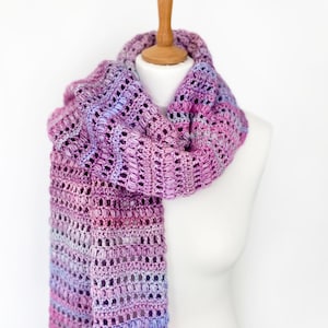 May include: A long, hand-crocheted scarf featuring a variegated yarn pattern in shades of purple, magenta, and light blue. The scarf displays a textured stitch pattern with alternating rows of openwork and dense, raised puff stitches throughout.