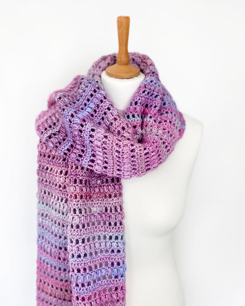 May include: A long, hand-crocheted scarf featuring a variegated yarn pattern in shades of purple, magenta, and light blue. The scarf displays a textured stitch pattern with alternating rows of openwork and dense, raised puff stitches throughout.