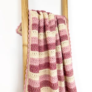 Può includere: Una coperta all'uncinetto a righe, fatta a mano, nei toni del rosa e del crema, drappeggiata su una scala di bambù. La coperta presenta un bordo smerlato e un motivo a punto strutturato. La combinazione di colori include strisce rosa chiaro e scuro.