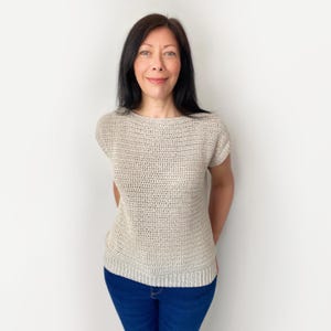 May include: A beige short-sleeved top featuring a textured crochet knit pattern with a boat neckline and a ribbed hemline, paired with dark blue denim jeans. The garment has a fitted silhouette and a consistent neutral colour throughout the knit.