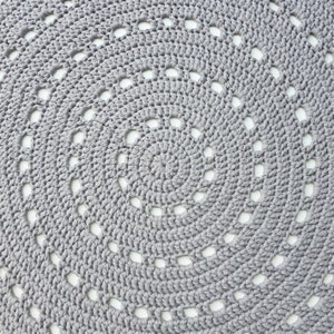 May include: A circular gray crochet mat featuring a repeating spiral pattern of thick yarn stitches. The design incorporates concentric rings of solid stitching interspersed with small, evenly spaced openwork gaps that create a textured effect.