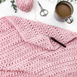 Pode incluir: Um cobertor de croch&ecirc; rosa claro com um padr&atilde;o texturizado, uma bola de l&atilde;, uma agulha de croch&ecirc; e uma tesoura. Uma caneca de caf&eacute; e galhos de vegeta&ccedil;&atilde;o tamb&eacute;m est&atilde;o presentes, sugerindo uma cena de artesanato aconchegante.