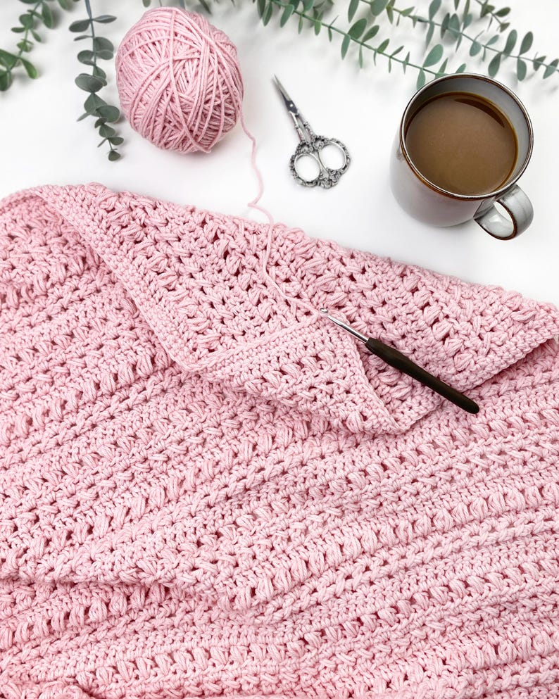 Pode incluir: Um cobertor de croch&ecirc; rosa claro com um padr&atilde;o texturizado, uma bola de l&atilde;, uma agulha de croch&ecirc; e uma tesoura. Uma caneca de caf&eacute; e galhos de vegeta&ccedil;&atilde;o tamb&eacute;m est&atilde;o presentes, sugerindo uma cena de artesanato aconchegante.