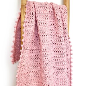 Pode incluir: Uma manta de croch&ecirc; rosa claro sobre uma escada de madeira. A manta apresenta um padr&atilde;o de ponto texturizado e uma borda ondulada. A manta feita &agrave; m&atilde;o &eacute; um item de decora&ccedil;&atilde;o aconchegante.