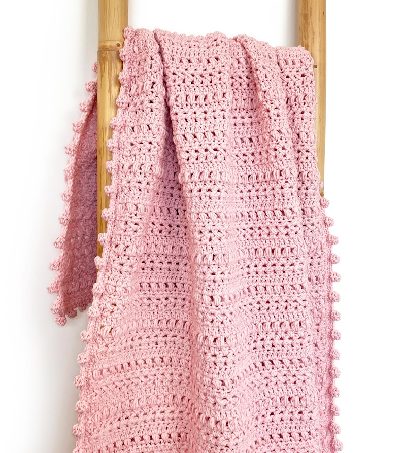 Pode incluir: Uma manta de croch&ecirc; rosa claro sobre uma escada de madeira. A manta apresenta um padr&atilde;o de ponto texturizado e uma borda ondulada. A manta feita &agrave; m&atilde;o &eacute; um item de decora&ccedil;&atilde;o aconchegante.