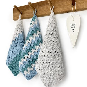 Op de afbeelding: Drie handgemaakte gehaakte washandjes in blauwe en grijze tinten hangen aan een houten rek. De washandjes hebben een textuur, open geweven ontwerp. Een wit hartvormig ornament met de tekst "All you need is love" hangt rechts.