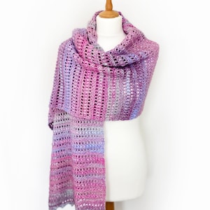 May include: A long, rectangular crochet scarf featuring a variegated color palette of magenta, purple, lavender, and soft blue. The textile displays a repeating pattern of vertical textured stitches and openwork rows, creating a lightweight, airy appearance.