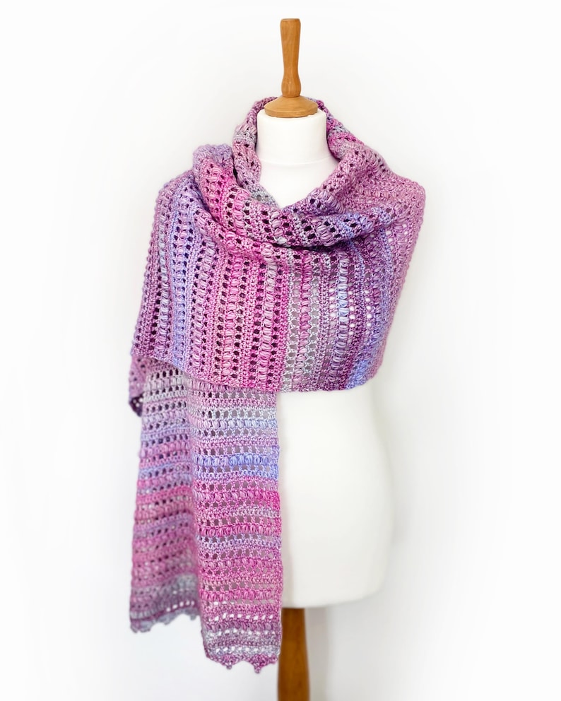 May include: A long, rectangular crochet scarf featuring a variegated color palette of magenta, purple, lavender, and soft blue. The textile displays a repeating pattern of vertical textured stitches and openwork rows, creating a lightweight, airy appearance.