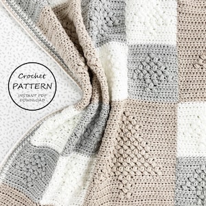 Puede incluir: Una manta de bebé tejida a crochet con un patrón de cuadros en beige, blanco y gris. La manta está hecha con un punto texturizado que crea una superficie elevada y abultada. La manta está doblada por la mitad y los bordes son visibles.