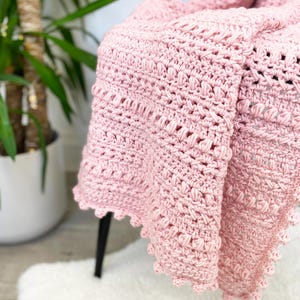 Pode incluir: Uma manta de croch&ecirc; rosa claro drapeada sobre uma cadeira. A manta apresenta um padr&atilde;o de ponto texturizado e uma borda ondulada. Esta manta macia e feita &agrave; m&atilde;o &eacute; um item de decora&ccedil;&atilde;o para casa.